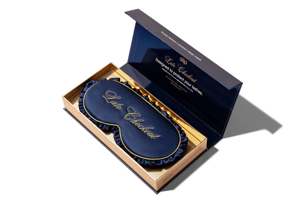 A luxury navy blue silk sleep mask with gold lettering, presented inside an open gold and navy blue gift box by Slip.